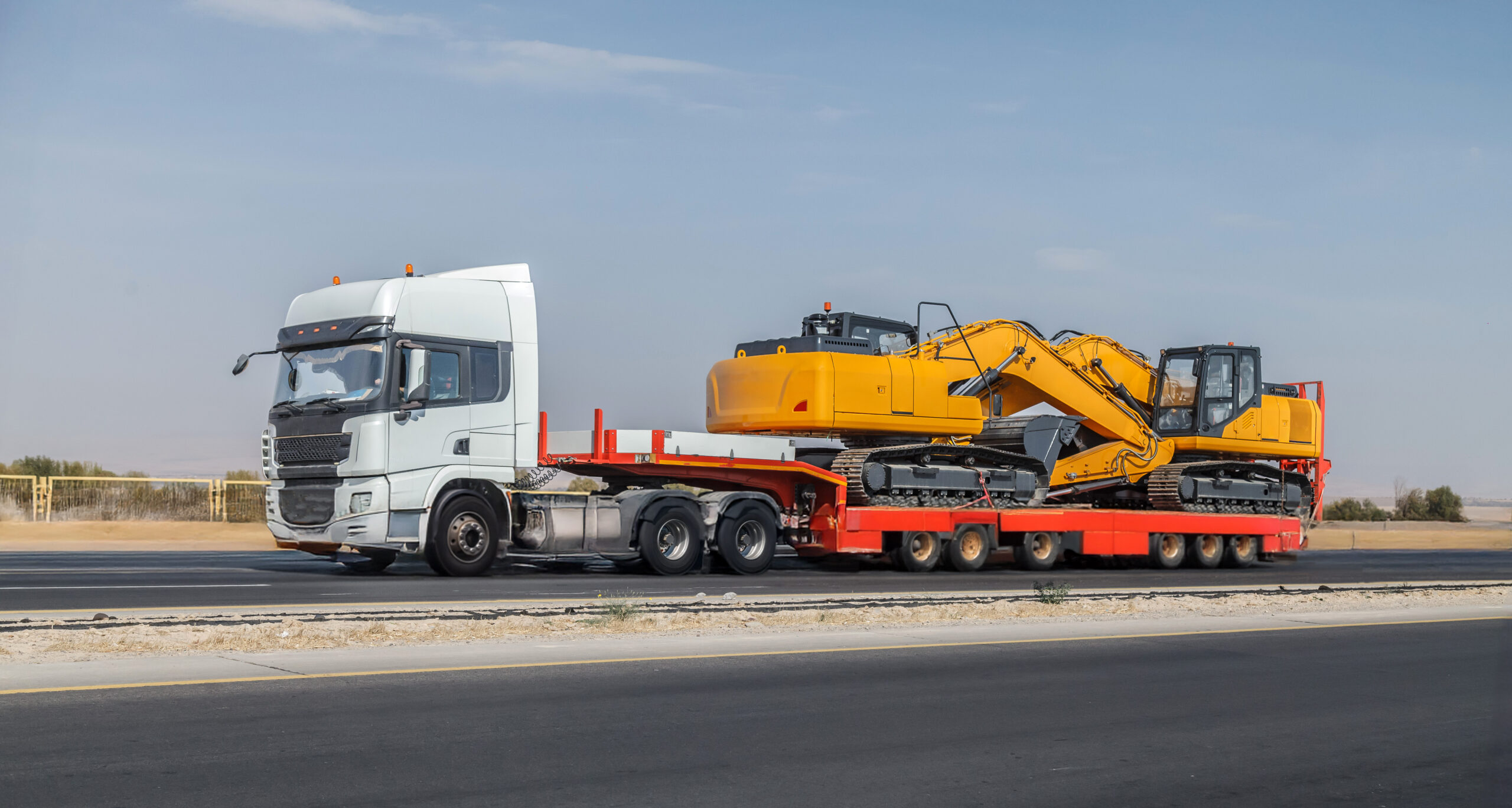 Large truck is hauling a trailer with a yellow and black machine on it. The truck is driving down a road, and the machine is securely strapped to the trailer